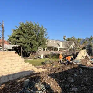 Construction site with a partially built wall and workers clearing debris under a clear sky.