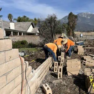 Workers building a brick wall outdoors with mountains in the background.